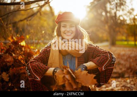 Bonjour l'automne. femme à la mode heureuse en chapeau rouge avec écharpe jetant des feuilles d'automne dans le parc de la ville. Banque D'Images