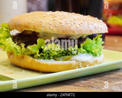 Un grand hamburger avec laitue et fromage sur une assiette verte. Le burger est garni de graines de sésame et de mayonnaise. Banque D'Images