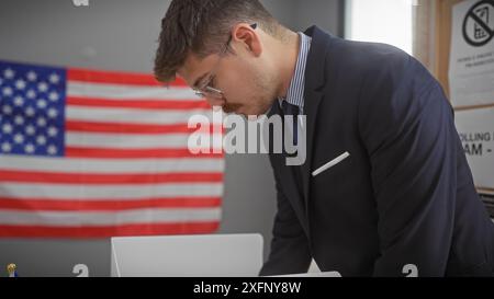 Un jeune homme d'affaires hispanique avec des lunettes et une moustache travaille sur un ordinateur portable dans un bureau à thème patriotique avec un drapeau américain. Banque D'Images