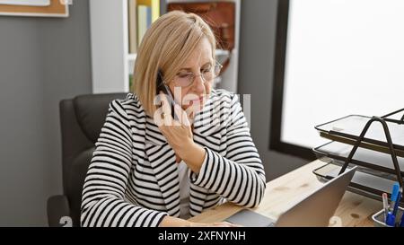 Femme d'affaires mature dans des lunettes parlant sur un smartphone dans un cadre de bureau moderne, montrant concentration et professionnalisme. Banque D'Images