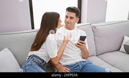 Un couple aimant profite d'un moment confortable ensemble à l'intérieur, avec la femme embrassant l'homme comme il tient un smartphone. Banque D'Images
