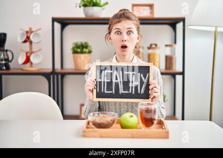 L'expression choquée traverse le visage de la jeune femme blonde à la maison, la peur et l'étonnement se mêlent alors qu'elle est assise avec son petit déjeuner sain et son tableau noir; Banque D'Images