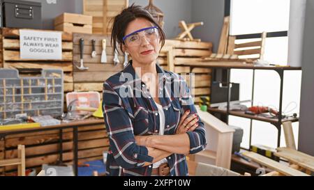 Femme mature confiante avec les bras croisés portant des lunettes de sécurité se tient dans un atelier de menuiserie. Banque D'Images