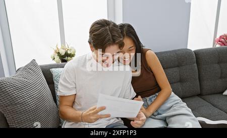 Un couple interracial heureux lisant un document ensemble sur un canapé dans un salon moderne, exprimant la joie et la convivialité. Banque D'Images