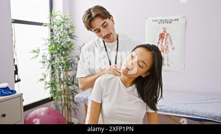 Un physiothérapeute masculin effectue une thérapie du cou sur une patiente dans une clinique avec un diagramme du système musculaire visible. Banque D'Images