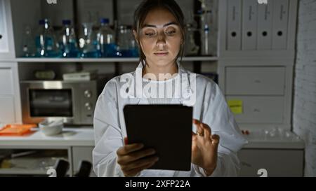 Femme hispanique scientifique utilisant la tablette en laboratoire avec fond de verrerie, reflétant l'intégration de la technologie de la santé. Banque D'Images