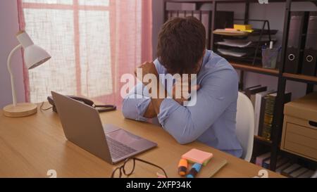 Un jeune homme hispanique tousse à son bureau de travail dans un bureau intérieur, entouré d'un ordinateur portable, d'une lampe et de fournitures de bureau. Banque D'Images