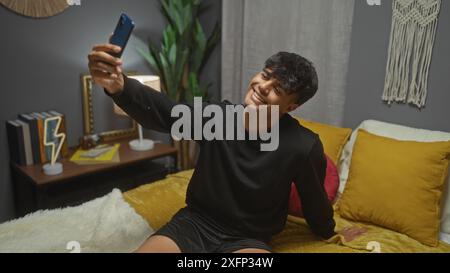 Jeune homme hispanique prenant un selfie dans une chambre confortable avec un décor moderne, y compris des livres, des plantes et des oreillers élégants. Banque D'Images