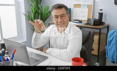 Un homme mûr en chemise blanche sourit dans son bureau, tenant une clé avec un ordinateur portable et des plantes d'intérieur derrière lui. Banque D'Images