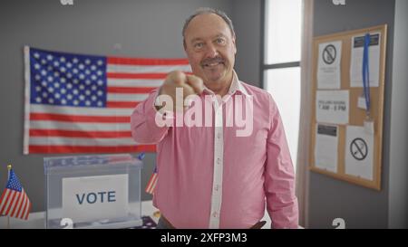 Homme d'âge moyen pointant la caméra dans un centre électoral américain avec drapeau américain et boîte de vote Banque D'Images