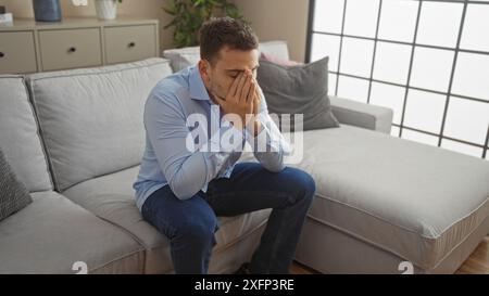 Un jeune homme hispanique avec une barbe est assis dans un salon se sentant stressé, le visage dans les mains, dans un environnement intérieur. Banque D'Images