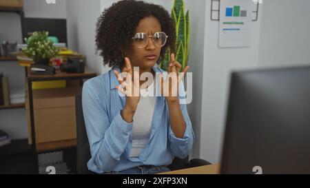 Une jeune femme afro-américaine aux cheveux bouclés croise les doigts en anticipation alors qu'elle est assise dans un environnement de bureau. Banque D'Images