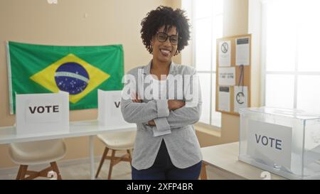 Une jeune femme afro-américaine aux cheveux bouclés se tient confiante avec les bras croisés dans une salle de collège électoral intérieure avec des drapeaux brésiliens et de la votine Banque D'Images