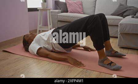 Une belle jeune femme afro-américaine effectue un exercice de bridge sur un tapis de yoga dans son salon, mettant l'accent sur sa routine d'entraînement ciblée dans un in Banque D'Images