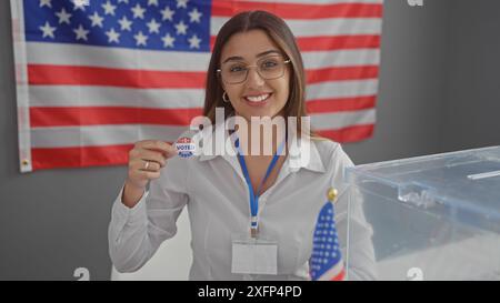 Jeune femme hispanique souriante montrant l'autocollant "j'ai voté" devant le drapeau américain dans un bureau de vote. Banque D'Images