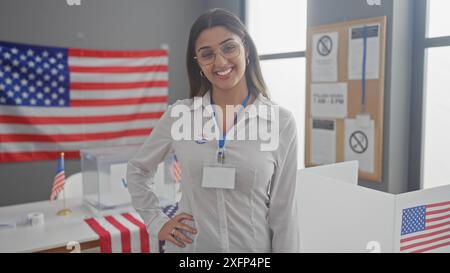 Jeune femme hispanique souriante avec un autocollant "j'ai voté", debout dans un bureau de vote avec un drapeau américain en arrière-plan Banque D'Images