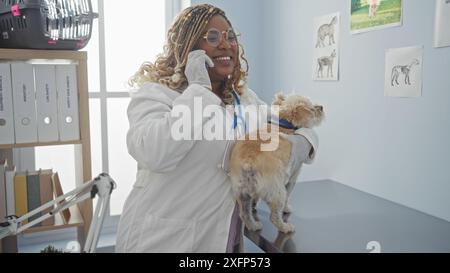 Une jeune femme afro-américaine vétérinaire avec des tresses, portant une blouse de laboratoire, tient un petit chien tout en parlant au téléphone dans une salle de clinique vétérinaire Banque D'Images