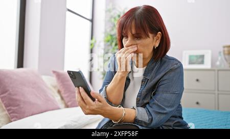 Femme âgée inquiète à l'aide d'un smartphone dans la chambre, montrant de la détresse alors que seul à la maison. Banque D'Images