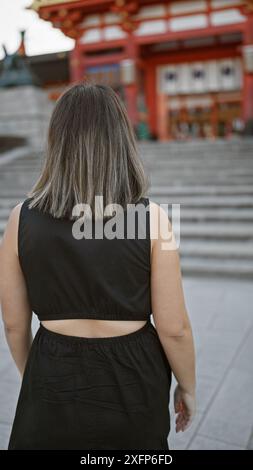 Vue arrière captivante d'une femme hispanique brune en lunettes, s'éloignant tranquillement, absorbée dans son smartphone, se promenant le long de fushimi inari taisha Banque D'Images