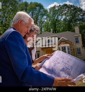 Couple senior examinant les plans de maison à l'extérieur de la maison de banlieue nouvellement construite, souriant et planifiant la future résidence dans le quartier résidentiel ensoleillé se Banque D'Images