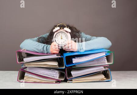 Femme fatiguée épuisée avec réveil dormant sur une pile de dossiers, burnout, stress et surmené, pression au travail Banque D'Images