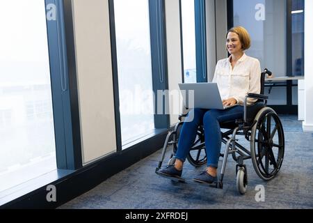 Femme souriante en fauteuil roulant à l'aide d'un ordinateur portable, travaillant dans un environnement de bureau moderne, espace de copie Banque D'Images