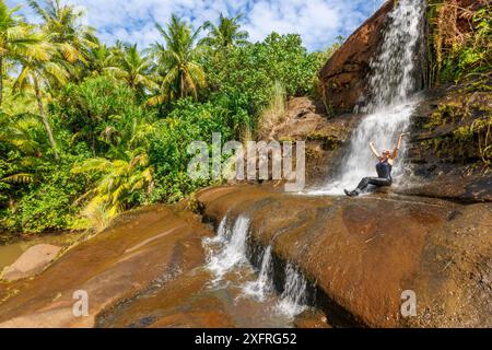 Une femme (MR) assise sous les chutes de Fintasa, avec des palmiers et un ciel bleu, Inarajan, Guam, Micronésie, territoire américain, océan Pacifique central. Banque D'Images