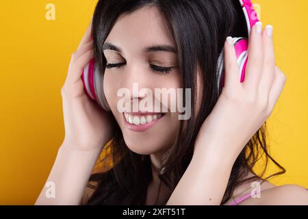 Jeune femme aux cheveux bruns sourit les yeux fermés, perdue dans la musique à travers des écouteurs roses sur fond jaune Banque D'Images