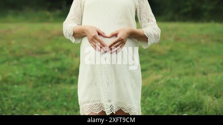 Grossesse, maternité, femme enceinte heureuse fait signe le cœur sur son ventre avec ses mains dans le parc d'été Banque D'Images