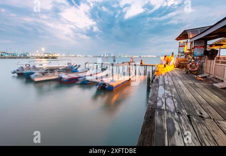 Penang Island, Malaisie-avril 25 2023 : le long de l'ancienne jetée promenades en bois, près du coucher du soleil, les visiteurs se tiennent debout, assis et prendre des photos au bord de l'eau, avec un petit moteur Banque D'Images