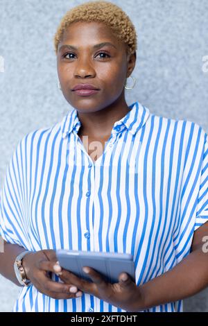 Tenant la tablette, femme afro-américaine dans la chemise rayée regardant réfléchi et concentré Banque D'Images