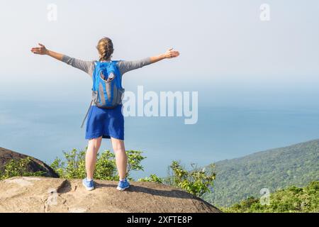 Jeune femme touriste levant la main et profitant de la vue sur la mer, l'air frais et le vent au sommet de la montagne. Fille avec sac à dos bleu. Banque D'Images