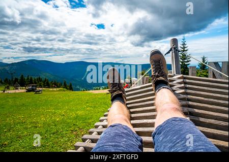 Randonneur de montagne se repose au bureau en bois au sommet de la colline. Vue du pied et des jambes du randonneur avec chaussures de trekking. Les collines dans loin, temps nuageux d'été. Banque D'Images