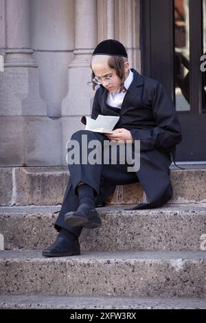 À la récréation, un jeune garçon juif orthodoxe studieux est assis sur les marches de l'école et étudie le Talmud. À Brooklyn, New York, mars 2024. Banque D'Images