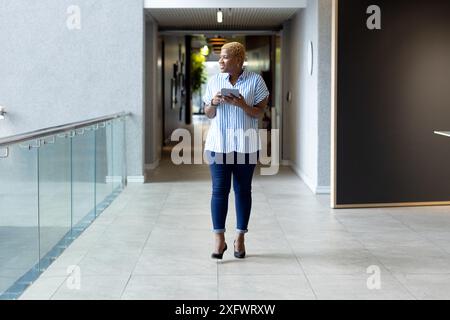 Tenant smartphone et regardant vers l'avant, femme afro-américaine marchant dans le couloir Banque D'Images