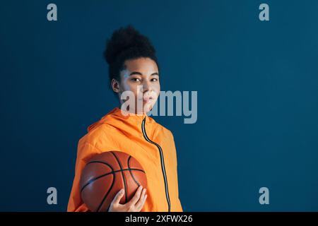 Jeune femme confiante avec un ballon de basket regardant la caméra. Femme portant une tenue de fitness orange tenant un basket-ball. Banque D'Images