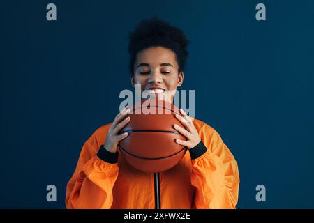 Femme avec basket portant des vêtements de sport orange. Femme souriante avec les yeux fermés tenant un ballon de basket. Banque D'Images