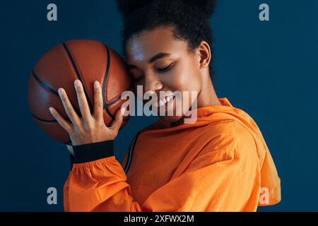 Gros plan d'une jeune femme souriante avec un ballon de basket. Femme avec les yeux fermés portant des vêtements de sport orange debout sur un fond bleu. Banque D'Images