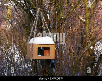 Un mangeoire à oiseaux bruns en bois et en contreplaqué recouvert de neige pend sur une corde sur les branches d'arbres en hiver. Banque D'Images