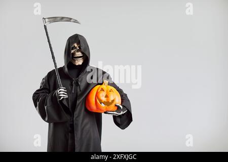 Homme en costume de mort de faucheuse sinistre isolé sur fond gris de studio tenant la citrouille et la faux Banque D'Images