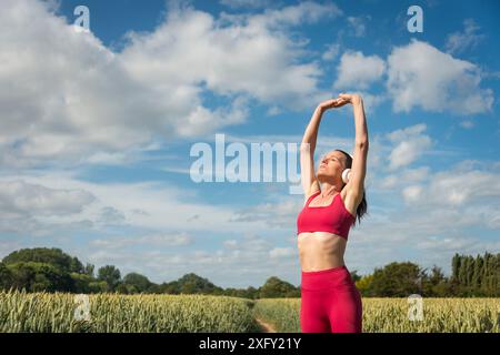 Femme sportive avec les bras levés faisant des exercices d'étirement dans le champ de blé, portant des vêtements de sport rouges avec un ciel bleu. Banque D'Images