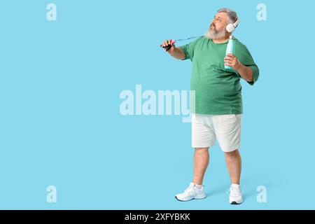 Homme mature en surpoids dans des écouteurs avec corde à sauter et bouteille d'eau sur fond bleu. Concept de perte de poids Banque D'Images