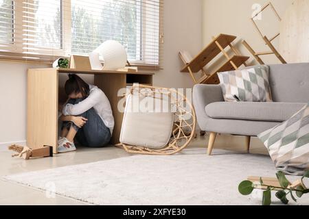 Jeune femme effrayée se cachant sous la table dans le salon pendant le tremblement de terre Banque D'Images