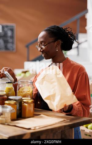 Cliente afro-américaine regardant un pot de pâtes tout en tenant un sac brun pour mettre des produits bio. Dame noire avec des lunettes examinant l'article fraîchement fait dans un récipient en verre au magasin local. Banque D'Images