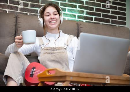 Heureuse jeune femme assise avec une tasse de café et ukulélé, portant des écouteurs par terre dans le salon de sa maison, regardant dans l'ordinateur portable Banque D'Images