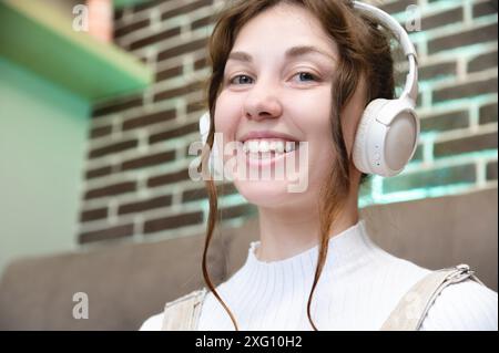 Portrait d'une jeune femme caucasienne souriante dans des écouteurs parlant par appel vidéo de la maison. Photo de femme heureuse dans le casque regardant la caméra ayant Banque D'Images
