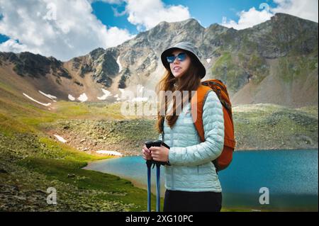 Une jeune femme caucasienne avec un sac à dos et dans un panama, se tient avec des bâtons de trekking près d'un lac de montagne et regarde au loin. randonnée, marche Banque D'Images