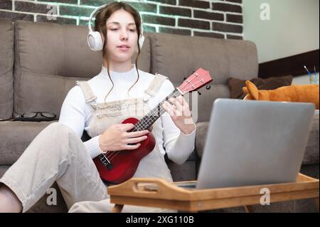 Une jeune belle femme est assise par terre à la maison et apprend à jouer de la guitare ou du ukulélé, debout à côté d'un ordinateur portable et d'une tasse de café. Banque D'Images