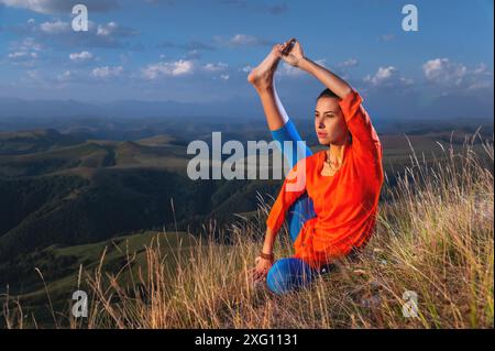 Jeune femme yogi attrayante pratiquant le yoga, faisant des variations d'exercices de Krounchasana, posture du héron, faisant de l'exercice dans la nature, portant des vêtements de sport Banque D'Images
