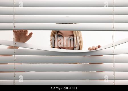 Jeune femme regardant à travers les stores de fenêtre sur fond blanc Banque D'Images